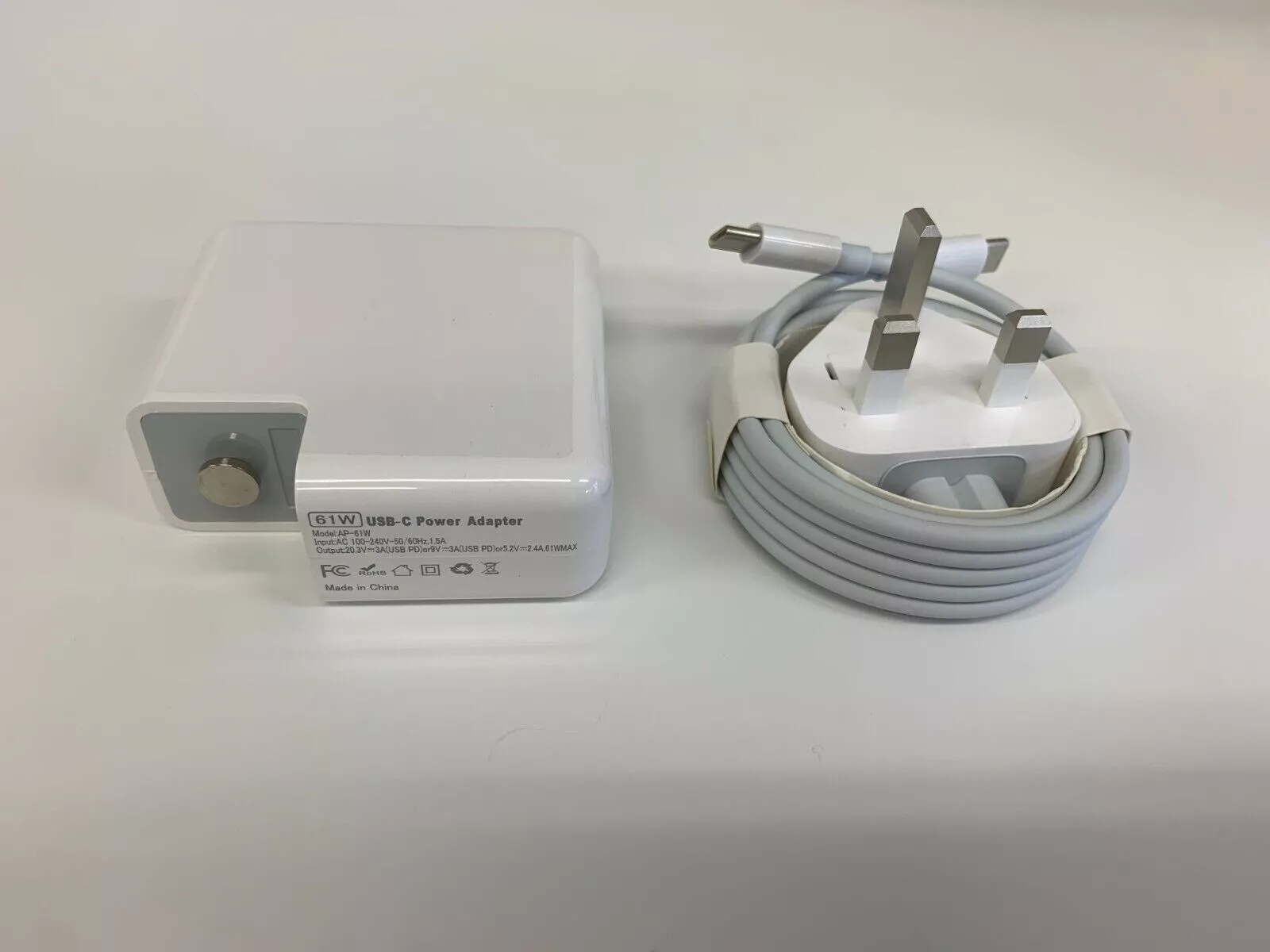 A 61W USB-C power adapter and coiled USB-C charging cable with a UK three-pin plug, included with the Apple MacBook Air 2020 M1 256GB/16GB, are placed on a white surface.