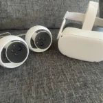 The Meta Quest 2 Virtual Reality Headset 128GB with Controllers, featuring a white headset with strap and two matching controllers with circular rings, rests on a gray fabric couch.