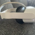 The Meta Quest 2 Virtual Reality Headset 128GB with Controllers, featuring a white design and fabric strap, sits on a grey couch with its indicator light on. A patterned cushion can be seen in the background.