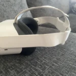 The Meta Quest 2 Virtual Reality Headset 128GB with Controllers, featuring a white design and fabric strap, rests on a gray fabric couch with a patterned cushion in the background.