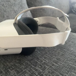 The Meta Quest 2 Virtual Reality Headset 128GB with Controllers, featuring a white design and fabric strap, rests on a gray fabric couch with a patterned cushion in the background.