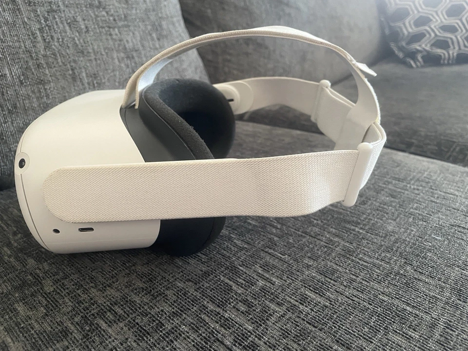 The Meta Quest 2 Virtual Reality Headset 128GB with Controllers, featuring a white design and fabric strap, rests on a gray fabric couch with a patterned cushion in the background.